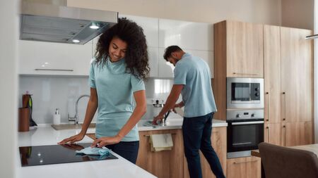 Clean Home. Two young professional cleaners in uniform working together in the kitchen. Happy afro american woman cleaning electric stove, young caucasian man wiping kitchen tableの写真素材