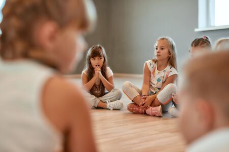 Having a break. A group of little cute girls sitting on the floor in the dance school. Choreography class. Little dancers sitting on the floor gathered around their female teacherの写真素材