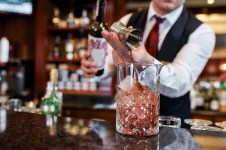 Young bartender standing at counter and pouring red wine to prepare iced cocktail in the bar. Horizontal shot. Selective focus on a drinkの写真素材