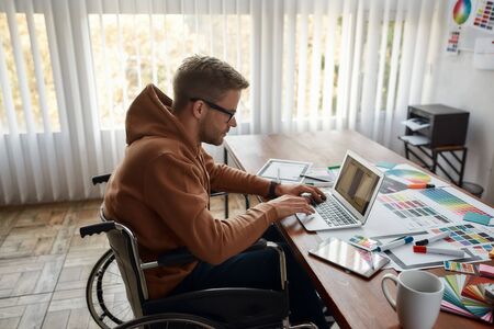 Creative professional. Side view of young male UX UI designer in a wheelchair designing new mobile app, working on laptop at his creative workplace in the modern office. User experience conceptの写真素材