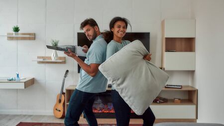 Having fun. Two young professional cleaners having fun while working together in the living room. Young caucasian man playing with handheld vacuum, afro american woman holding sofa pillow and smilingの写真素材