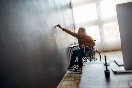 Working on presentation. Young male professional designer in a wheelchair writing something on big blackboard in the modern office. Portrait of disabled young man sketching some ideasの写真素材