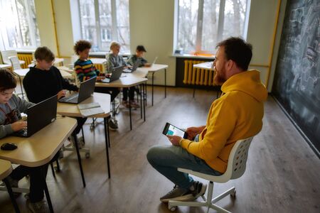 Where Learning Begins. Portrait of young male teacher communicating with his pupils, while sitting with tablet pc near the blackboard during a lesson in modern smart schoolの写真素材