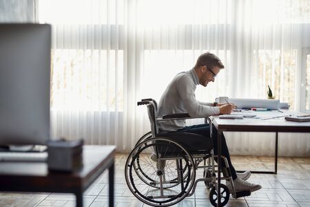 Great working day. Side view of cheerful male office worker in a wheelchair drawing something and...