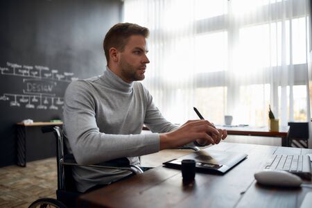 Being creative. Side view of young designer in a wheelchair using stylus pen and digital tablet while sitting at his workplace in the modern office. Design studioの写真素材