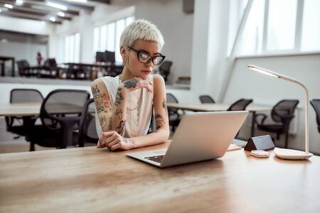 Busy day. Portrait of focused blonde tattooed businesswoman in eyeglasses looking at laptop screen while working in the modern working spaceの写真素材