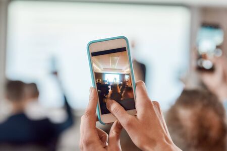 Get more out of now. Back view of participant hands holding smartphone while taking picture, video on corporate business meeting at the conference hallの写真素材