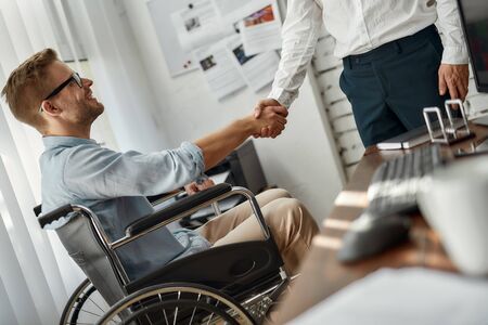 Cropped view of male office worker in a wheelchair shaking hand to his colleague while working together in the creative office. Teamwork. Disability and handicap concept. Business meetingの写真素材