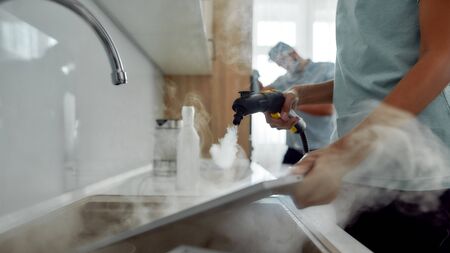 Dirty work. Cropped photo of a woman in uniform cleaning filters for kitchen range hood with steam cleaner while her male colleague working on the backgroundの写真素材