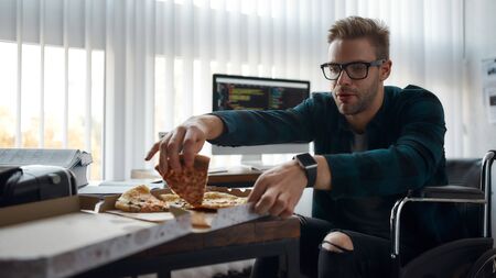 Relaxing at work. Young male web developer in a wheelchair eating pizza while sitting at his...