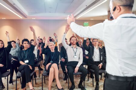 Be prepared. Audience listening to asian male speaker and raising hands to ask a question at the conference hall. Man answering questions while giving a talk at corporate business meeting, forumの写真素材