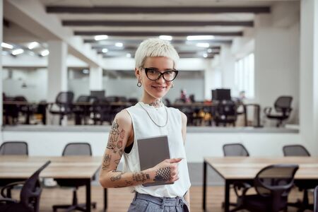 Great working day. Portrait of pretty tattooed blonde businesswoman in eyeglasses holding digital tablet and smiling at camera while standing in the modern officeの写真素材