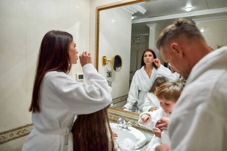 But first, hygiene. Caucasian parents and two kids in white bathrobes having morning routine in hotel bathroom, brushing teeth while standing near the mirror. Family, travel, resort, vacation conceptの写真素材