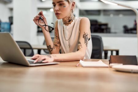 Focused at work. Young and beautiful tattooed woman typing something on the laptop while sitting at her working place in the modern officeの写真素材