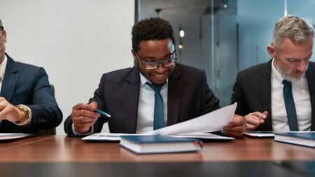Great result. Young african businessman in formal wear analyzing some documents and smiling while sitting at the office table in the modern officeの写真素材