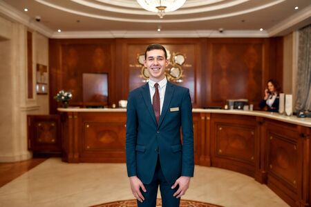 Ready to help. Happy male receptionist worker in uniform looking at camera with a smile while standing at hotel lobbyの写真素材