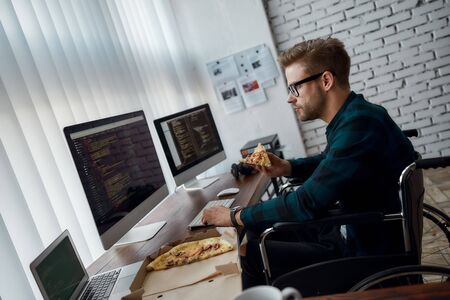 Focused male web developer in a wheelchair eating pizza and writing program code while sitting at...