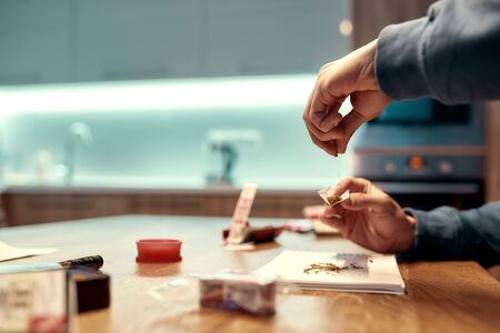 Close up of male hands making a marijuana cigarette or joint at home in the kitchen. Marijuana tools on the table. Cannabis and weed legalization concept. Selective focus. Side view. Horizontal shotの写真素材