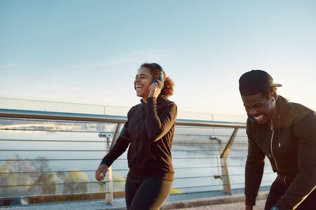 Sport that brings happiness. Young happy african couple in sportswear and headphones jogging together on the bridge in the early morning, they are laughing and enjoying runの写真素材
