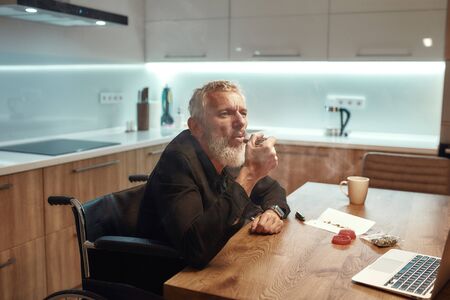 Time to think. Senior disabled man in wheelchair smoking marijuana from a metal pipe, sitting in the kitchen. Marijuana tools on the table. Legalization and dependence on light drugs conceptの写真素材