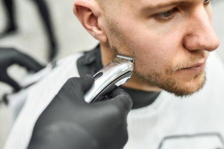 Close up photo of professional barber in black gloves trimming beard of his client with electric clipper machine. Shaving, grooming, beard correction. Mens haircut. Barber equipmentの写真素材