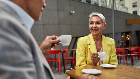 Perfect morning. Beautiful and cheerful middle-aged woman in yellow suit jacket drinking fresh coffee with her friend while sitting in cafe outdoorsの写真素材