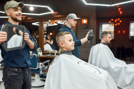 Haircut for little boy. Young smiling barber holding a mirror and showing the result of his work to cute little boy sitting in barber shop chair. Child visiting barber shopの写真素材
