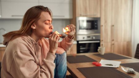 Reducing Stress. Young caucasian woman lighting cannabis in the bowl of metal pipe while sitting in the kitchen. Buds in a plastic bag and red marijuana grinder on the tableの写真素材