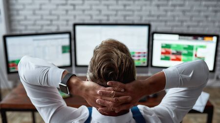 Rear view of middle-aged trader sitting by desk in front of pc and analyzing stock market chart, while working in the office. Stock trading, people, business concept. Horizontal shotの写真素材