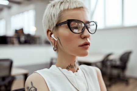 Enjoying music at work. Portrait of young blonde tattooed business woman in eyeglasses, with short haircut wearing wireless earphones while working in the modern officeの写真素材