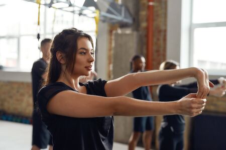 Make yourself proud. Portrait of young woman in black sportswear looking focused, stretching arms while working out in industrial gym. Group training conceptの写真素材