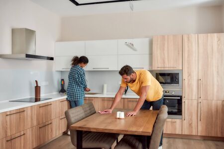 Lets work together. Young couple in casual clothes cleaning kitchen together. Caucasian man cleaning kitchen table, while afro american woman washing dishes on the backgroundの写真素材