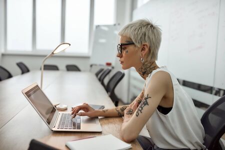 Businesswoman focused at work. Side view of young and beautiful tattooed woman with short haircut using laptop while sitting at her working place in the meeting roomの写真素材
