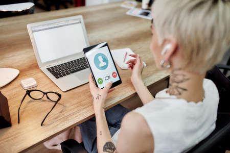 Video conference. Back view of young blonde tattooed business woman using her touchpad, having video call while sitting at her working place in the office. Digital technologiesの写真素材