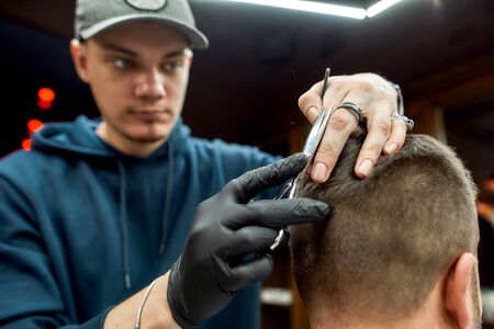Working process. Young barber in cap with scissors in hands making haircut for his client in barber...