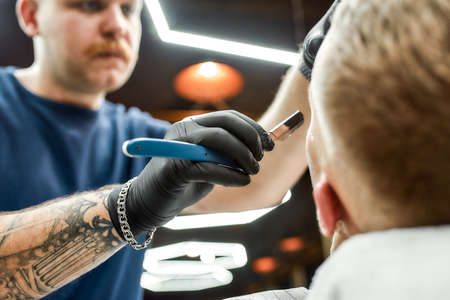 Beard correction. Male barber in black gloves with tattooed arm shaving client with dangerous...