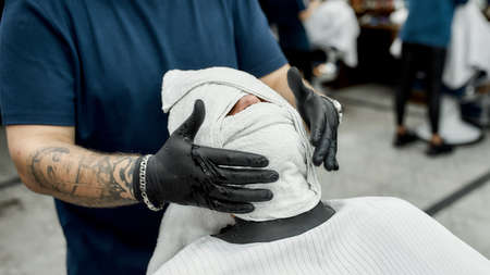 Professional tattooed barber covering face of his client with hot towel, steaming face before...