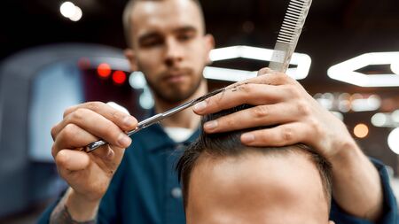 Personal stylist. Close up of young professional barber with scissors and hair comb in hands doing haircut for his client in barber shopの写真素材