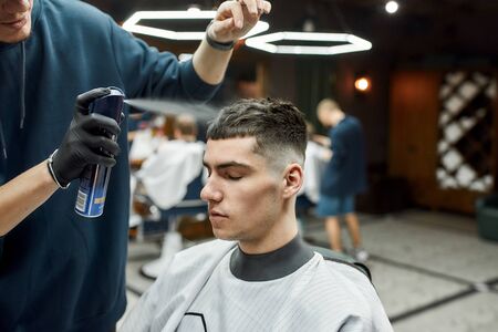 Barber fixing haircut of his client with hair spray while working in the barbershop. Young man visiting barbershop. Selective focusの写真素材