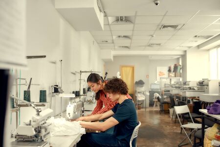 Careful measures. Young man, clothing designer sitting and sewing. His female colleague helping him. Dressmaker working on the sewing machine. Tailor making a garment in his workplaceの写真素材