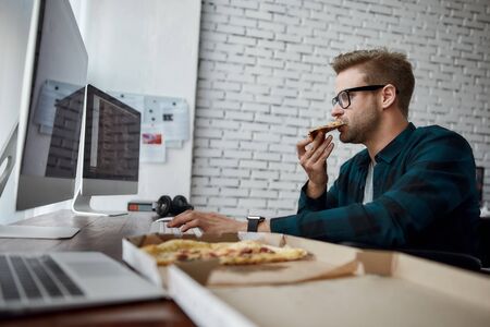 Feeling hungry. Side view of hungry male web developer in a wheelchair eating pizza and working on desktop computer while sitting at his workplace in the modern officeの写真素材