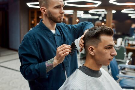 Professional barber with tattoo on his arm holding comb and making hairstyling for young handsome guy who is sitting in a barbershop chairの写真素材