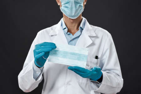 Cropped photo of a male doctor in medical uniform and blue sterile gloves showing medical mask at camera while standing against black backgroundの写真素材