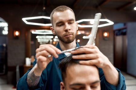 Working in barbershop. Close up of young professional barber with scissors and hair comb in hands making haircut for his client in barber shopの写真素材