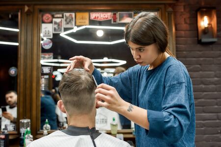 Cool hairstyle. Young caucasian man getting new haircut in the modern barbershop. Professional barber girl doing haircut for her client. Beauty salonの写真素材