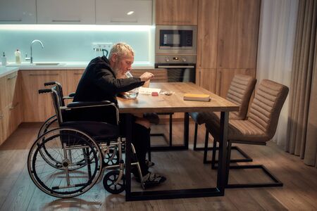 Painkiller. Senior disabled man in wheelchair lighting cannabis in the bowl of glass water pipe or bong in kitchen. Marijuana tools on the table. Legalization and dependence on light drugs conceptの写真素材