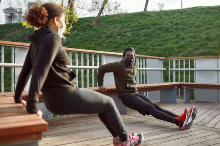 Social distancing. African couple in protective masks working out in park outdoors. Doing push-ups exercises on bench, training tricepsの写真素材