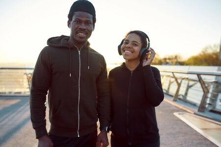 Cheerful healthy african couple after run. Positive male and female runners in headphones standing on the bridge in the morning and smiling at cameraの写真素材