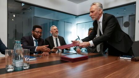Presenting new project. Mature businessman in formal wear giving folders to his multicultural colleagues while having a meeting in the officeの写真素材
