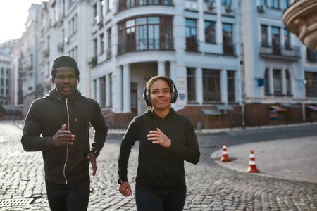 Keep moving. Happy young sporty African couple in headphones jogging, running together through the city street in the early morningの写真素材
